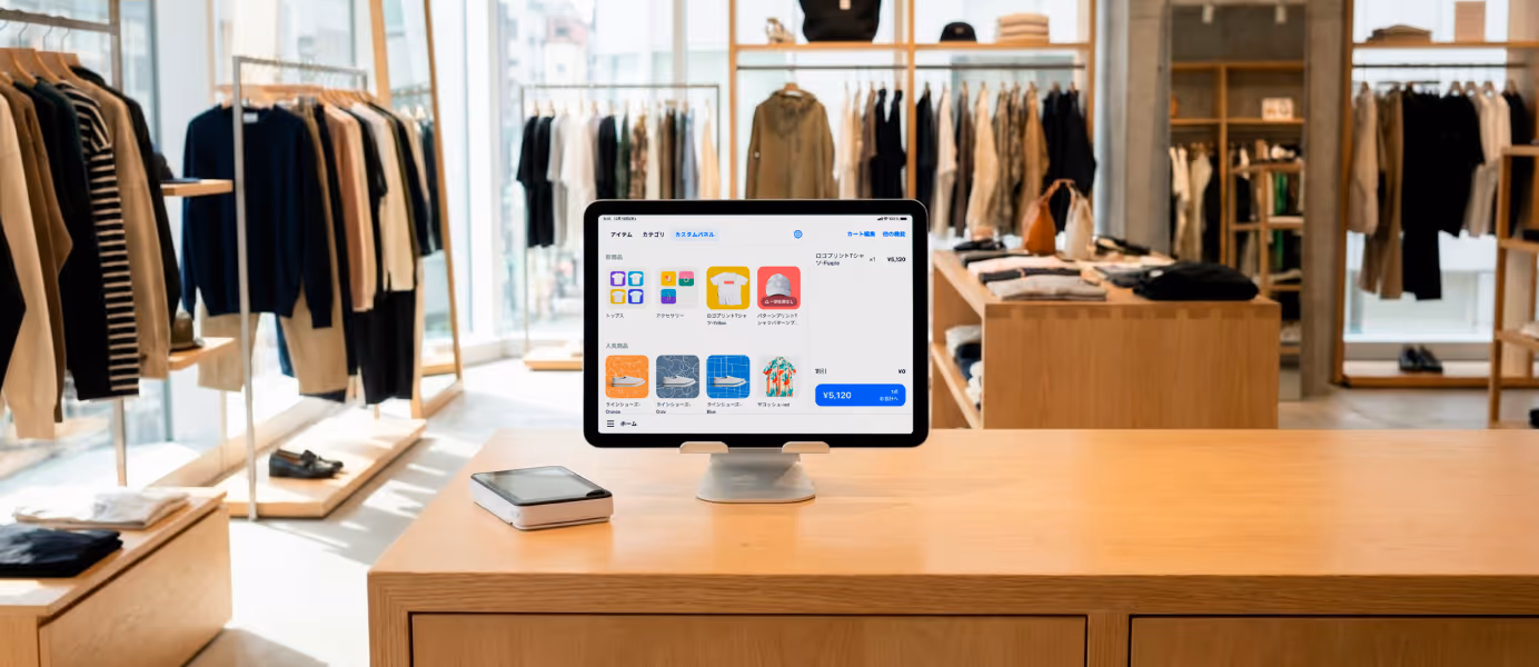 洋服店のカウンターに置かれたPOSレジアプリの画面を表示したタブレット端末と決済端末。背景にはさまざまな衣類が並ぶ店内。
